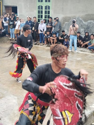 This image depicts an outdoor cultural performance in the city of Kerjo, Indonesia.
In the foreground, a male performer, appears to be in motion, his upper body angled to the right. He has light brown hair with a patterned headband and facial markings including red and black streaks around his eyes and smudges on his forehead and nose. He wears a black long-sleeved shirt, a red sash, and patterned black, white, and red lower garments. He holds a red, white, black, and yellow stylized horse head prop with black horsehair-like features, gripping it with both hands.
Behind him and slightly to the left, a second male performer is visible. He shares similar facial markings and attire, including a black long-sleeved shirt with a logo, black pants with decorative yellow and red fabric near the ankles, and a black, white, and red checkered skirt-like garment. He wears black and white sneakers and holds an identical stylized horse head prop.
In the background, a crowd of approximately 30-40 spectators, consisting of adults, teenagers, and some children, observes the performance. They are positioned along a low concrete wall or ledge, some standing, some seated. Their attire is casual. One individual on the right, wearing a light brown jacket, light blue jeans, and a black face covering, holds a smartphone or camera device horizontally, aiming it towards the performers. Another individual in the mid-left background also holds a phone, appearing to record the event.
The performance takes place on an uneven, wet concrete or paved surface. Behind the spectators is a plain, light grey concrete building wall featuring a light green wooden window frame with multiple panes in the center, and an open doorway to the far left. The overall lighting is diffuse, suggesting an overcast sky.