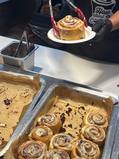 A close-up shot captures a person in black gloves using red-tipped tongs to place a frosted cinnamon roll onto a white paper plate. The cinnamon roll is golden brown with a swirl of frosting on top. In the background, two aluminum trays filled with more cinnamon rolls are visible. The tray in the foreground is partially filled, with some cinnamon rolls already removed, leaving behind a messy trail of frosting and chocolate sauce on the parchment paper. A metal container with tongs is also visible in the upper left corner. The overall scene suggests a food service environment, likely a bakery or cafe, where cinnamon rolls are being prepared or served. The location of the scene in Shanghai, China, is not discernible from the provided image.
