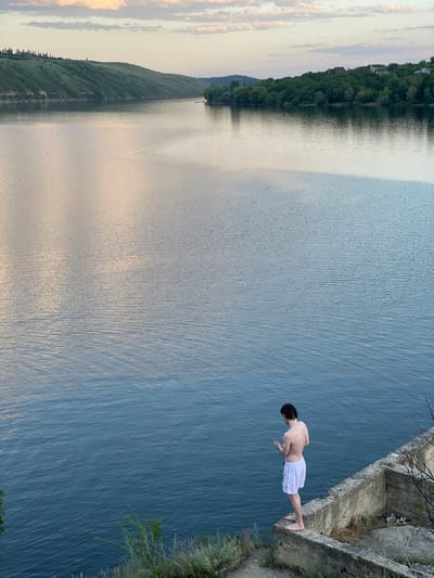 A man in white shorts stands on the edge of a high cliff, preparing to jump into a large body of water. The cliff is constructed of concrete and stone, with some vegetation growing around its base. The water appears deep and stretches out to the horizon where green hills are visible in the distance. The sky is mostly clear with some light clouds. The image is rotated on its side.