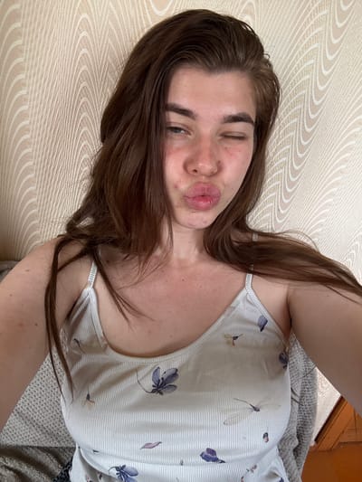 A young woman with long, brown hair is pictured from the chest up, winking and puckering her lips. She is wearing a light-colored, ribbed tank top with a floral pattern. The background is a beige, textured wallpaper with a wave-like design. The woman appears to be indoors, possibly in a residential setting. There is no visible indication of the city of Мещёры, Russia.