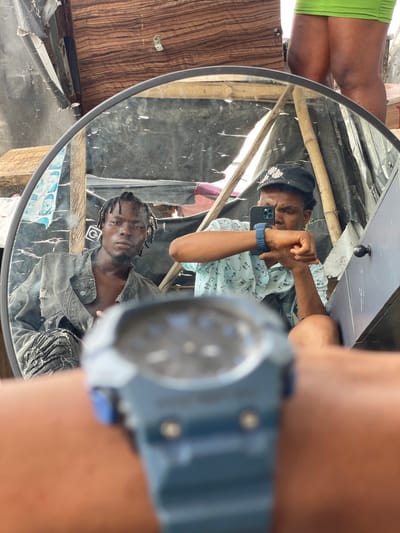 In Ojomu, Nigeria, a close-up shot shows a mirror reflecting two young men and a portion of a woman's legs. The man on the left, wearing a distressed denim jacket and ripped jeans, looks directly at the camera with a serious expression. His hair is styled in dreadlocks. The man on the right, wearing a patterned shirt and a black cap, is holding a smartphone and appears to be taking a selfie. He has a blue watch on his wrist. In the foreground, a blurred blue wristwatch is visible, suggesting the photographer is wearing it. The background is cluttered, with wooden planks, bamboo poles, and tarpaulin, indicating a possibly informal or construction-adjacent environment. A woman's tanned legs, clad in a bright green mini-skirt, are visible in the upper right portion of the frame, suggesting she is standing behind the photographer.