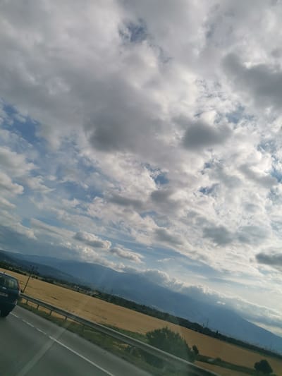 The image is a perspective shot taken from a vehicle moving along a road, possibly a highway, in the vicinity of Kazichene, Bulgaria. The foreground shows a portion of the road with white lane markings and a metal guardrail. To the right of the road, a field of golden crops, likely wheat or barley, stretches towards a range of blue-hued mountains in the background. The mountains are partially obscured by a hazy atmosphere and appear to be densely forested at their base. A dark-colored vehicle, possibly an SUV, is visible in the lower left quadrant of the image, indicating movement. The sky is filled with a dramatic display of clouds, ranging from fluffy white cumulus to darker, more ominous formations, interspersed with patches of blue sky. The overall scene suggests a rural or semi-rural landscape under a dynamic sky, possibly captured during a journey through the Bulgarian countryside near Kazichene.