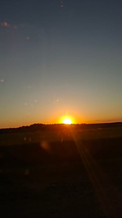 The image shows a sunset in Любанскі сельскі Савет, Belarus. The sun is setting on the horizon, with orange light. The foreground consists of dark land or foliage, while the sky transitions from dark blue to orange. There are some spots of light or lens flares scattered in the sky.