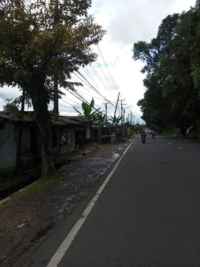 The scene depicts a roadside environment under an overcast sky, with a paved asphalt road dominating the midground and extending into the background. The location cannot be visually confirmed as Serpang Shalli, Bhutan from the image content.
On the left side of the road, an unpaved, dark, wet, and uneven dirt/gravel shoulder is present, bordered by a narrow concrete drainage ditch. A large tree with green-brown foliage stands beside the ditch. Behind the tree and ditch are multiple low-rise, rudimentary structures with corrugated metal roofs, the first of which appears light blue/grey. A red plastic bag hangs near an entrance of one structure. One individual, wearing dark clothing, is visible on the dirty shoulder near these structures, appearing to be sweeping or engaged in ground-level work. Further along this side, a dark fence or wall is visible, behind which green vegetation, including large-leafed plants resembling banana trees, grows. Numerous utility poles with a dense network of black electrical or communication lines crisscross the sky above these structures.
The asphalt road is dark grey, marked with a continuous white line on its left edge and intermittent yellow dashed lines in its center further down. Two individuals are operating motorcycles on the road, traveling away from the camera; the lead rider is wearing a dark helmet and jacket. Additional vehicles or figures are faintly visible in the far distance on the road.
On the right side of the road, dense dark green trees line the edge, extending into the background. Distant hills or mountains are faintly discernible through atmospheric haze in the far background. The sky is overcast with white and grey clouds.