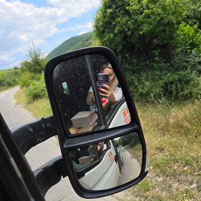 In the side mirror of a white truck, a blonde woman with a purple phone takes a selfie. She is wearing a white t-shirt and a red top underneath. A small brown box is visible in the truck's cabin. The truck is driving on a paved road that curves through a rural, green landscape. The sky is blue with some clouds. A red and white sticker is affixed to the side of the truck. The scene appears to be in or near the city of Peshtera, Bulgaria.