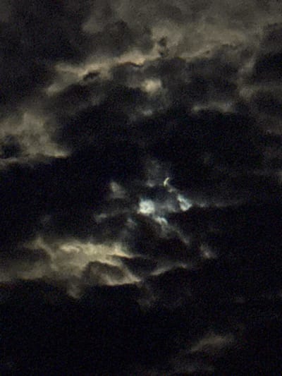 The image is a dark, grainy, and abstract representation of what appears to be a night sky. It is dominated by deep black and very dark grey tones, interspersed with irregular, blurry patches of white and light grey. These lighter areas suggest dense clouds or atmospheric formations, partially illuminated by an unseen light source, possibly the moon, which might be subtly hinted at by a faint, crescent-like shape visible in the central region of the original image and some crops. The significant amount of visual noise and grain throughout the image indicates photography under very low light conditions, typical of nighttime. There are no discernible people, distinct objects, or specific activities depicted. No text is visible. The scene portrays cloudy weather at night. The location context provided for the image is Strazhitsa, Bulgaria, though no specific geographical features are discernible within the frame itself.