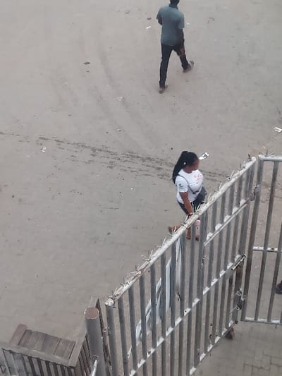 The scene, located in Agani, Nigeria, is an elevated view of a dusty, light-colored ground surface with scattered debris and faint tire tracks. Two individuals are present. In the upper frame, a male subject, viewed from the back, walks leftward, wearing a dark short-sleeved shirt, dark trousers, and light-colored shoes. In the middle frame, a female subject walks rightward, adjacent to a metal barrier. She has dark hair in a ponytail, wears a white short-sleeved t-shirt, dark lower garment, and light-colored sandals, carrying a dark shoulder bag. The metal barrier, with vertical bars and anti-climb spikes along its top, occupies the lower right. A wooden step structure is visible in the bottom-left corner.