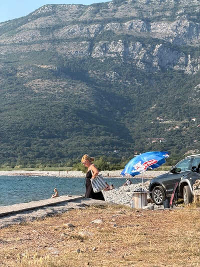 This media file captures a sun-drenched scene along the coast of Buljarica, Montenegro. A woman, dressed in a black dress and carrying a striped tote bag, stands on a rocky, grassy embankment. She is looking down towards the clear blue water, possibly preparing to enter or having just emerged from it. A blue and white umbrella, bearing the text "Frikom" and an image of a cartoon character, is open nearby, providing shade. Behind the umbrella, a dark-colored car is parked. The background is dominated by a steep, densely forested mountain, characteristic of the Montenegrin coastline. The water is calm, reflecting the bright sunlight, suggesting it's a warm, clear day. The overall atmosphere is one of leisure and relaxation, indicative of a holiday or vacation.