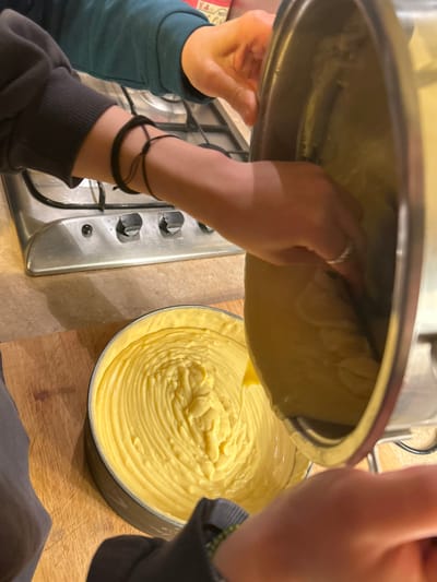 Two individuals pour yellow batter from a stainless-steel pot into a round baking pan, already partially filled. One person wears a ring and a dark-colored bracelet. A stovetop with silver knobs is adjacent to the pan. The scene is set on a wooden countertop. The angle suggests an overhead shot captured at close range.