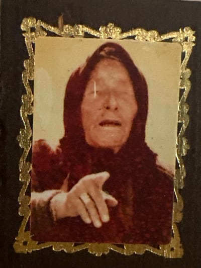 A framed portrait of an elderly person wearing a dark headscarf and a dark garment. The person has their face obscured by a vertical white line, but their mouth is open and their right hand is pointing forward, with the index finger extended. The portrait is set against a dark background and is surrounded by a gold, ornate frame with a raised, decorative pattern. The image appears to be a photograph. There is no indication of the city of Marten, Bulgaria, in the image.