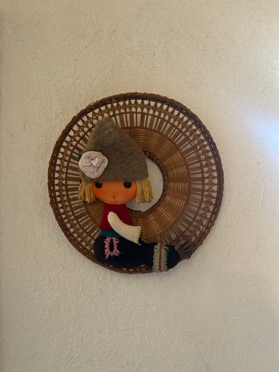 A handmade textile doll with a pointed brown hat adorned with a light pink rose is affixed to a round, woven wicker frame. The doll has a round orange face with black button eyes and small red dots for a mouth. Yellow yarn hair extends from beneath the hat. The doll wears a red shirt, a white sleeve, and black pants with a green trim and a pink embroidered detail. The doll's lower body appears to be shaped like a mermaid's tail, with a brown, pleated fin on the right side. The wicker frame is circular with an intricate woven pattern and is attached to a textured, off-white wall. The overall composition is a decorative wall hanging. The location in Parvomay, Bulgaria, is not discernible from the image.