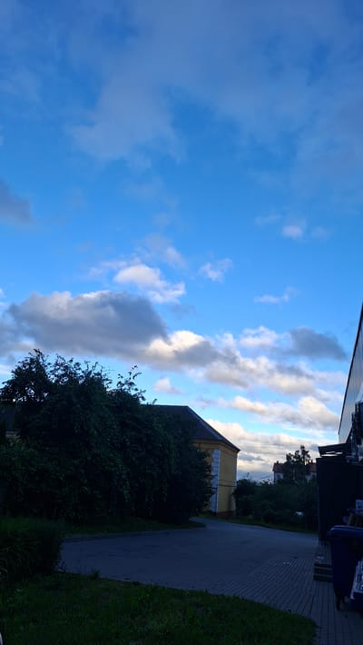 The image shows an outdoor scene with a blue sky filled with scattered white and grey clouds. In the foreground, there is a paved area with a dark asphalt surface and grey cobblestones on the right side. Lush green grass is visible along the bottom left of the frame. To the left, a dense, dark green bush or tree dominates the mid-ground, partially obscuring a yellow building with white trim. The building appears to be an older, perhaps institutional structure, with a dark roof. To the right, a modern, dark-sided building is partially visible, with some industrial-looking equipment or ventilation units mounted on its exterior. Two large blue trash bins are situated near the edge of the paved area on the far right. In the distance, beyond the yellow building, a few other structures and trees can be seen. The overall impression is that of an urban or semi-urban environment, possibly a commercial or institutional area, under a partly cloudy sky. The specific location in the city of Smalyavichy, Belarus is not explicitly identifiable from visual cues within the image itself.