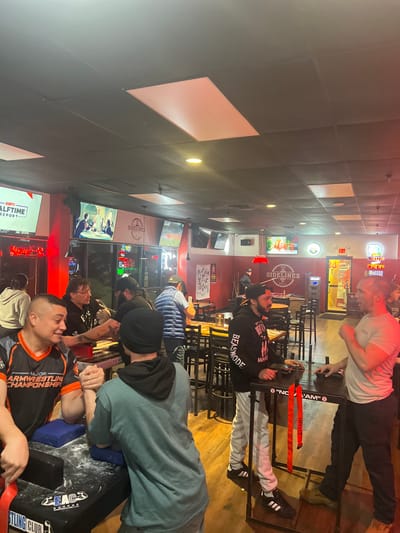 Inside a brightly lit establishment, possibly a sports bar, several individuals are engaged in arm wrestling matches. The scene includes multiple arm wrestling tables set up, with participants facing off. Televisions displaying "SIDELINES" and "ESPN HALFTIME REPORT" are visible in the background. Signage references "MAJOR ARMRESTLING CHAMPIONSHIP," indicating an organized event. Participants wear casual attire, and the atmosphere appears lively and competitive.