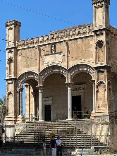 This image depicts the facade of a stone building, likely a church or historical monument, under a clear blue sky. The building exhibits classical architectural elements such as arched entrances, columns, and ornate stonework. A wrought-iron fence runs along the front of the building, partially obscuring the entrance and a set of stone stairs leading up to it.
Several people are visible in the scene. Two individuals stand near the entrance, one appearing to be indoors or at the threshold of a dark opening. Another group of people are positioned further back, near the top of the stairs, with one person looking towards the camera. On the right side of the image, a person is seated or leaning near the bottom of the stairs, and another is walking away from the camera. The presence of people suggests the location is open to visitors.
The sunlight casts shadows, indicating it is likely daytime, possibly late morning or afternoon. The clear sky suggests fair weather. The architecture and the context of Palermo, Italy, point to a historical site. There is some visible text on a plaque on the building, but it is too small and indistinct to be read. The overall mood is one of peaceful exploration of a historical landmark.