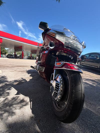 A low-angle, close-up shot captures the front of a dark red Honda motorcycle parked on asphalt in Sotonići, Montenegro. The motorcycle is the primary subject, positioned prominently in the foreground, with its windshield reflecting the blue sky and sparse white clouds. A red and white gas station canopy is visible in the background, with several cars at the pumps. Trees and mountainous terrain are also present, suggesting an outdoor, possibly rural or semi-rural, location. The scene is illuminated by bright sunlight, casting distinct shadows of the motorcycle and surrounding foliage onto the ground. There are no people clearly visible in the frame.