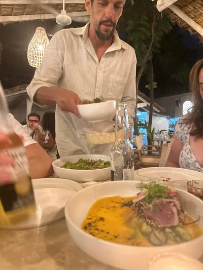 A group of people are seated at an outdoor dining table at night. The location appears to be a restaurant in Brisas de Zicatela, Mexico, evidenced by the beach setting and architecture. A man with a beard is wearing a striped shirt, holding a plate, and seemingly engaged in a conversation with other people at the table. A woman in a floral-patterned dress is partially visible near the top of the frame. Various dishes, including a salad and a tuna dish with mango and avocado, along with water bottles, are arranged on the table. Another person is visible wearing a mask near the bottom of the frame. The lighting includes a combination of natural light from the surroundings and artificial lighting from a ceiling fan and decorative lamps.