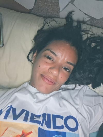 A woman with dark, curly hair lies on her back, facing the camera. She is wearing a white t-shirt with blue lettering that reads "VIEMICO" and a graphic image. Her head is slightly tilted to her left. She has a nose ring and an earring in her visible ear. The background shows a light-colored pillow and a tiled surface, suggesting an indoor setting. The lighting is dim, and the image appears to be a selfie. This scene is located in El Valle del Espíritu Santo, Venezuela.