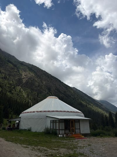 In a mountainous region of Kyrgyzstan, a traditional yurt, a round, portable dwelling, stands prominently in the foreground. The yurt has a white exterior with a decorative red and white pattern around its upper section. A covered porch with windows extends from one side of the yurt, and a set of yellow steps leads to the entrance. Several people are visible on the porch, appearing to be gathered or seated. The yurt is situated on a dirt path, with green grass and vegetation surrounding it. In the background, a steep, tree-covered mountain rises, with a dramatic sky filled with large, white clouds above. The overall scene depicts a serene, natural landscape with a cultural dwelling.