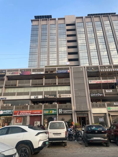 This media file captures a street scene in Ahmedabad, India, with a prominent modern office building dominating the frame. The building features a glass facade reflecting the sky and surrounding environment. On the ground level, several businesses are visible, including "Parshwa Dental Care & Implant Center," "NNTOURS," "insights 10," "SBI Securities," "ICICI Bank," "BUDDHA LUXURIOUS SPA," and "FUSION PIZZA." Other text includes "Salé & Pepe," "PINKY'S," "BIG BURGER CO.," and "hoggeterlans Multi Cuisine Restaurant." Multiple vehicles, including cars and motorcycles, are parked along the street, suggesting it's a busy area. The scene appears to be set during the daytime, with clear skies. There are no prominent individuals or specific activities highlighted, but the presence of businesses and vehicles implies a typical urban commercial environment. License plates visible on cars read "GJ27BE5897" and "GJ27ED4200".