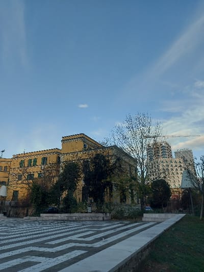 The image, presented sideways, captures an urban scene under a wide expanse of clear blue sky with scattered, wispy white clouds, which dominates the left half of the frame. Towards the center and right, an architectural landscape unfolds.
In the mid-right, a traditional-style, multi-story building with a light yellow facade and visible green shutters on its windows stands, partially obscured by the bare branches of trees. Green ivy or climbing plants cling to parts of its walls. Further in the upper right background, a taller, modern high-rise building with a distinctive white geometric pattern or texture on its exterior is visible, and a construction crane extends upwards near its top.
In the lower right quadrant, a patterned paved pathway featuring a repeating labyrinth-like or geometric design in white lines on a darker surface stretches diagonally across the frame. Adjacent to this pathway, a narrow strip of green grass or ground cover runs along its upper edge.
The scene appears to be set during daytime, likely with ample sunlight given the bright sky. The bare trees suggest either a late autumn or winter season. There are no people visible, nor any discernible activities taking place. No text is visible in the image.