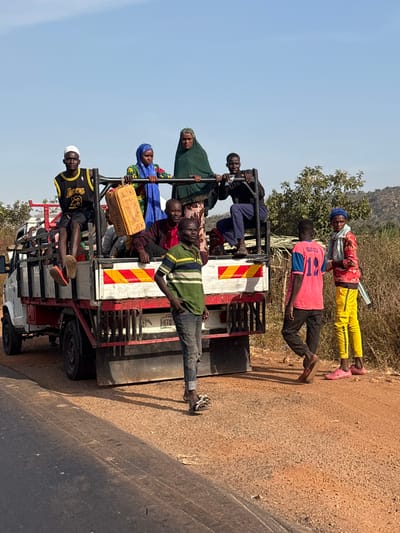 This image captures a white flatbed truck stopped on a dirt road shoulder adjacent to a paved road in Kwakwa Laji, Nigeria. Approximately seven to eight individuals occupy the truck's bed, which has red-framed railings and a white and orange chevron reflective strip on the tailgate. A young male in a white skull cap, black "LAKERS 23" tank top, and black shorts sits on the left railing, holding a yellow plastic jerrycan. Standing centrally in the truck bed are two women: one in a blue head covering and a patterned dress, and another in a dark green head covering and a patterned skirt. On the right railing, a male in a dark blue long-sleeved shirt and pants is seated. Other individuals are partially visible within the truck bed. Three males stand on the dirt ground beside the truck: one centrally located in a green and blue striped shirt, dark jeans, and dark sandals; another to his right in a pink shirt bearing "ESAN CTU 12" and dark pants; and a third on the far right, wearing a light blue patterned head covering, red long-sleeved shirt, bright yellow pants, and pink sandals, holding a white stick. The environment features dry, sparse vegetation and small trees on the right, with distant hills visible under a clear, bright blue sky in the background.
