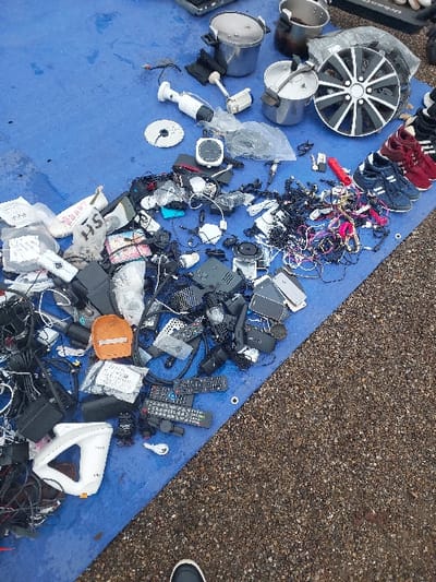 This image depicts an outdoor market stall in Eckbolsheim, France. A large blue tarp is spread across a gravel or crushed stone ground surface, serving as a display for a variety of used items.
In the upper right portion of the tarp, three stainless steel pots, two of which appear to be pressure cookers, are arranged. Adjacent to these is a multi-spoke car wheel, predominantly black with silver accents. To the right of the wheel, several pairs of athletic shoes, including dark blue and red varieties, are lined up. A white cylindrical security camera and a white immersion blender are also visible in this area.
The central and left sections of the tarp are densely covered with miscellaneous electronic components and accessories. This includes extensive piles of tangled black and multicolored cables, power adapters, and numerous remote controls of various designs. Small black electronic boxes, potentially routers or media players, are scattered throughout these piles, some individually and others grouped. An orange, padded protective case is visible among the electronics. In the lower left quadrant, the hemispherical base of a white Apple iMac G4 computer is identifiable. Many smaller items, including various connectors and components, are enclosed in clear plastic bags.
At the bottom center of the frame, the toe of a human foot, wearing a dark shoe with a white sole, is partially visible, indicating a person's presence near the display. The scene is illuminated by natural daylight.