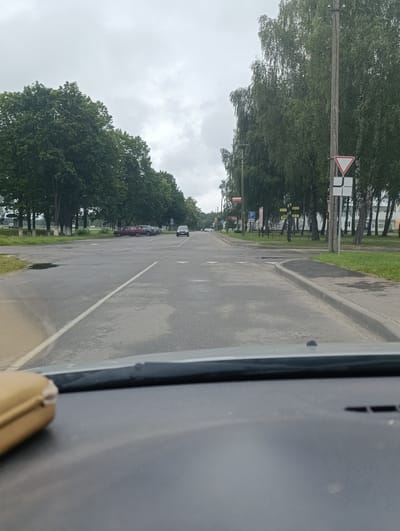 The image shows a view from inside a vehicle driving on a wet asphalt road in Ленінскі сельскі Савет, Belarus. The road is lined with trees on both sides, with a cloudy sky overhead. There are several cars parked on the right side of the road and some buildings in the background. Traffic signs are visible, including a yield sign and a parking sign. The interior of the car, including the dashboard and windshield, is partially visible in the foreground.