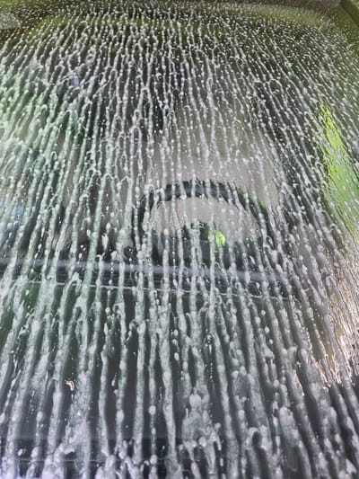 The image shows a close-up view of a window covered in soap suds and water streaks, suggesting it is being washed or has recently been washed. The suds create horizontal patterns across the glass. Through the soapy window, a blurred, dark background is visible, with hints of green from vegetation outside and vertical lines that might indicate blinds or a fence. The lighting suggests it is daytime, possibly overcast or in diffused light, as there are no harsh shadows and the light is even across the surface of the glass. The scene is abstract and textural, focusing on the patterns created by the water and soap on the glass rather than a clear depiction of the outside world. There are no people, animals, or specific objects identifiable beyond the window itself and the suds. The location context of Issy-les-Moulineaux, France, does not provide any direct visual cues within the image itself.