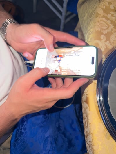 A person holds a smartphone with a green case in their hands. The screen shows a photograph of a street in Florence, Italy. The street appears to be narrow and lined with buildings. A few people are walking along the street. The background includes a table with a yellow cloth with floral patterns and a blue cloth with floral patterns. The person's wrists are visible, and they are wearing a watch.