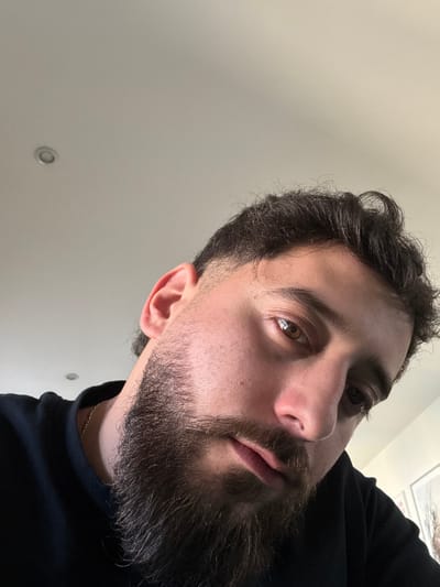 A close-up, low-angle photograph taken in Lingolsheim, France, depicts an adult male looking directly towards the camera. The subject possesses dark brown, wavy hair, styled with a faded cut on the sides, and a full, dark brown beard. His visible right eye features a brown iris. He is dressed in a black round-neck garment, and a thin gold-colored chain is visible around his neck. The background comprises a plain, light-colored ceiling, with a single recessed light fixture positioned in the upper left quadrant of the frame. A faint outline suggesting a framed piece of art or photograph is discernible on a wall in the lower right background. The scene is uniformly and brightly lit, indicative of an indoor setting.
