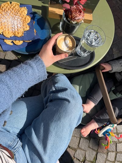 The image depicts a scene at an outdoor cafe in Meisenheim, Germany. The table is a light green color and holds a glass of water, a cappuccino in a paper cup held by a person wearing a blue sweater and jeans, a vase with a pink rose, a black serving tray, a menu card, and a blue plate with a waffle dusted with powdered sugar on a blue napkin. A child is seated in a stroller, visible by their legs, feet, hands, and a colorful toy attached to the stroller. The ground is paved with cobblestones.