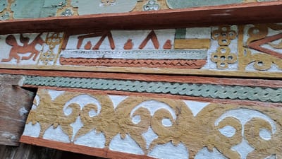 This media file showcases a close-up view of ornate wooden carvings, likely part of a traditional building in Meunasahmeucap, Indonesia. The carvings are layered and painted in earthy tones of orange, yellow, and green, with white as a background.

The top layer features a frieze with stylized motifs. On the left, a reddish-orange figure with wing-like protrusions resembles a mythological creature or a stylized horse. To its right, a geometric pattern of interlocking circles in yellow stands out, followed by a section depicting what could be abstract representations of boats or structures, rendered in yellow and reddish-orange.

Below this, a band of green carved wood displays a repeating, intricate pattern of interconnected oval shapes. Further down, another carved wooden element is visible, featuring elaborate scrolling patterns in a textured yellow, set against a white background, bordered by reddish-orange wood. A piece of weathered, unpainted wood is visible on the left, suggesting the material's age and exposure.

The overall impression is of decorative architectural elements, rich in cultural artistry. The details of the carving and painting suggest a traditional craft. There is no visible text in the image. The lighting suggests it was taken during the daytime.