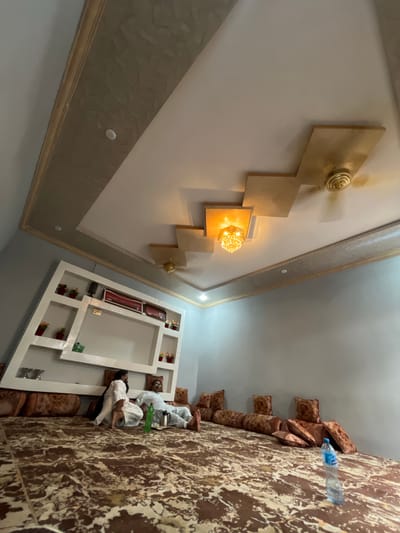 The image depicts the interior of a room, possibly located in Zhob, Pakistan. A ceiling fixture with geometric gold-toned accents and integrated lighting is prominently displayed on the ceiling. The walls are painted white. The floor shows patterned and textured fabric, likely part of a couch or seating arrangement. Two individuals, wearing white clothing, appear to be lying down, possibly resting or sleeping. A plastic bottle is visible near the edge of the fabric. A white shelving unit or decorative structure is attached to the wall. The overall environment seems to be a domestic setting within a building.
