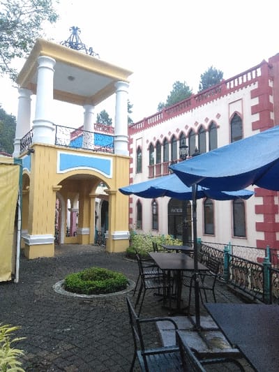 The image depicts an outdoor courtyard scene within the Pueblito Antiguo area of Parque de Diversiones, located in La Uruca, Ciudad de San José, Costa Rica.
To the left, a prominent two-story, yellow and white structure with blue accents stands. Its lower level features arched passageways. The upper level is an open pavilion supported by four white columns, topped with a yellow cornice and a dark, ornate finial. A black decorative metal railing surrounds the upper platform.
To the right, a multi-story building with a distinctive architecture is visible. It features a light cream facade with red trim outlining arched windows and defining decorative elements along the roofline, which includes crenellations. The windows are tall and arched. A sign displaying "PATISSE" is affixed to one of the windows. A decorative black metal railing separates this building from the courtyard's seating area.
In the mid-ground, an outdoor café seating area is present, featuring multiple dark rectangular tables and matching chairs, all unoccupied. Two large blue patio umbrellas are open, providing shade. The ground is paved with dark grey interlocking pavers. Centrally located within the courtyard is a circular raised garden bed containing green shrubbery.
Sparse green foliage and trees are visible in the background against an overcast, bright sky. No individuals are present in the scene, and no specific actions or interactions are depicted.