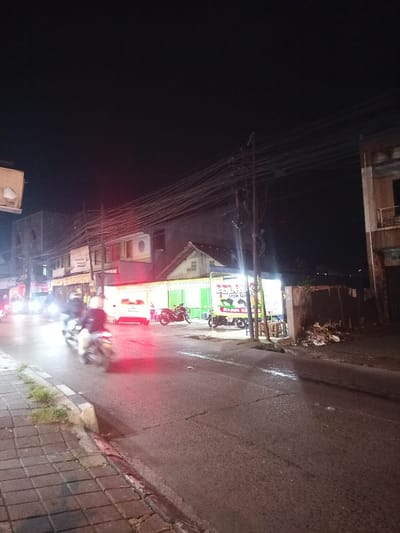 This image depicts a nighttime street scene in Ciomas, Indonesia. The view is from a tiled sidewalk on the left, looking down a two-lane asphalt road that appears wet in areas and shows signs of cracking.

Numerous overhead utility lines span horizontally across the upper middle of the frame, connected to at least two dark utility poles on the right side of the road. On the left, a series of multi-story buildings line the street, featuring unreadable signage and an exposed air conditioning unit on one facade.

Traffic is present on the road. Multiple motorcycles are in motion on the left side, exhibiting motion blur and illuminated headlights. One rider is clearly visible on a motorcycle in the foreground, wearing a dark jacket and helmet. A white sedan with illuminated red taillights is visible further down the road, facing away from the camera.

On the right side of the road, a brightly illuminated food stall is set up under a temporary structure, featuring a banner with Indonesian text, including "TELOR AYAM BAK" and "AYAM KECAP." Chairs or stools are visible in front of the stall. Two motorcycles are parked near the stall. Adjacent to the food stall and behind a low wall, a pile of assorted trash is visible on the ground. A two-story building with exposed concrete is visible on the far right. The overall illumination is provided by artificial sources, including vehicle lights and the food stall's lighting. The sky is dark and featureless.