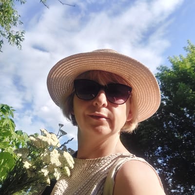 In Voronkiv, Ukraine, a woman with short blonde hair, wearing a straw hat and sunglasses, poses for a selfie outdoors. She is holding a bouquet of small white flowers. The background consists of a bright blue sky with scattered clouds and lush green trees. The woman is wearing a textured, light-colored sleeveless top. The image captures a moment of outdoor leisure and enjoyment in a natural setting.