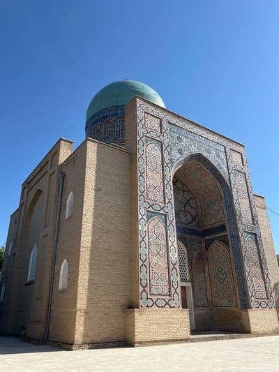 This is a low-angle, eye-level outdoor shot of a building in Samarkand City, Uzbekistan, under a clear blue sky. The structure is primarily made of light brown brick. The main visible facade features a large, ornate archway decorated with intricate blue, turquoise, and pink tilework, forming geometric patterns and Arabic calligraphy. Above the arch, the building continues with more tiled decoration and a large turquoise dome, also adorned with blue tilework. The left side of the building has a flat brick wall with arched windows that have white latticework. The ground in front of the building is paved with light-colored square tiles. There are no people or animals visible in the image. The architectural style suggests a historical religious or governmental building, consistent with the prevalent styles in Samarkand.
