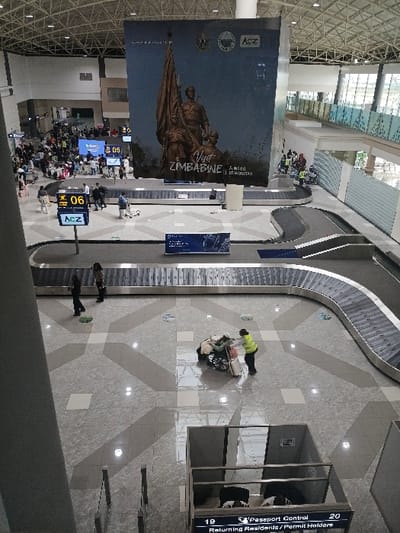 This image displays an elevated view of the baggage claim and arrivals hall at Robert Gabriel Mugabe International Airport in Harare, Zimbabwe. The facility is modern and spacious, featuring a high, vaulted ceiling with exposed white structural trusses and a gray and white geometric tiled floor.

Three curving baggage carousels are prominent. In the central foreground, an individual wearing a yellow-green high-visibility vest pushes a luggage cart laden with several bags and a wheelchair. Two other individuals walk in the midground near a carousel. Approximately 30-50 people are present in the background, congregated around additional carousels and walkways, indicative of passengers or staff.

A large vertical banner hangs centrally from the ceiling, depicting a bronze statue, the text "Visit ZIMBABWE," and the "AcZ" (Airports Company of Zimbabwe) logo. Digital displays are visible, including one showing "06" and "AcZ" near a carousel, and another in the bottom right corner indicating "Passport Control" with numbers "19" and "20" for "Returning Residents/Permit Holders." The area is brightly lit and appears fully operational.