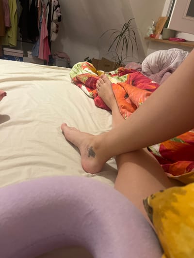 The photograph shows a human subject lying in a bed. The bed has a white sheet and a floral-patterned blanket. A lavender-colored neck pillow is visible. The subject has bare legs and feet, with a turtle tattoo on one ankle. Background includes a shelf with various items, a potted plant, and clothing. The location is an attic room. The city of Saarbrücken, Germany, cannot be determined from the content of this image.
