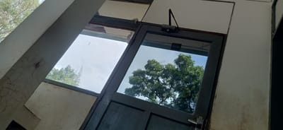 The media file depicts a section of a building's exterior, focusing on a large, dark-colored door with a glass panel and a smaller, adjacent window. The door is a double door and has a silver handle. Above the door, a hydraulic door closer is visible. The glass panel of the door reflects the sky and lush green trees, suggesting an outdoor setting. The window to the left also offers a view of trees and the sky. The building's structure is visible, with parts of the walls painted in a light, off-white color. One of these walls shows signs of cracking and wear. The time of day appears to be daytime, with bright natural light and a clear sky, indicating fair weather. The scene is likely an entrance or part of a building in a tropical or subtropical area, given the foliage. There is no visible text in the media file.