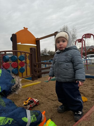 A young child wearing a gray hooded jacket and beige knit hat is standing in a sand-filled playground at a location in Akulovo (Gp Kubinka), Russia. The child is looking directly at the camera. Behind the child is a playground structure with a wooden frame, an orange and yellow sail-shaped element, and a blue and red tic-tac-toe game. In the sand around the child's feet are various toys, including a yellow shovel, a toy car, and other plastic items. Another child in a blue patterned jacket is partially visible in the lower right corner of the frame. The sky is overcast.