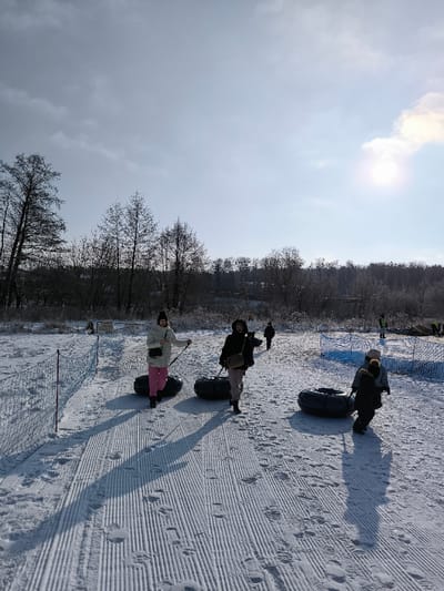 The image depicts a winter scene at a snow tubing area in Prybuzke, Ukraine, on a bright, sunny day. The foreground shows a snow-covered slope with distinct, parallel groomed tracks, indicating a path for walking uphill while pulling snow tubes. Numerous footprints are visible on these tracks.
In the immediate foreground, a total of five individuals are visible, progressing uphill from left to right.
* On the left, a woman wearing a white puffer jacket, pink pants, and a dark beanie is pulling a black snow tube by its rope. She holds a light-colored object in her left hand, possibly a phone or a small bottle.
* To her right, slightly behind, a second woman in a dark jacket, light-colored pants, and a dark beanie is also pulling a black snow tube. Her face is partially obscured by shadow.
* Further to the right and slightly behind, a child in a dark jacket is pulling a third black snow tube. An adult figure, largely obscured, walks behind or alongside this child.
* In the mid-ground, two additional figures are seen walking uphill, one of whom is distinct in a white jacket and dark pants, and another smaller figure behind them.
To the left of the groomed path, a low orange safety net barrier runs along the edge of the snow. Further right, a blue netting also appears to delineate a section of the snow. The background features a dense line of bare deciduous trees and some coniferous trees, suggesting a forest or treeline bordering the recreational area. In the far background, faint structures are visible amidst the trees. The sky is bright blue with scattered white clouds, and the sun, positioned in the upper right quadrant, casts long, prominent shadows from the individuals onto the snow, indicating a low sun angle typical of winter.