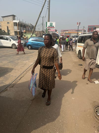 The image captures an outdoor street scene in a commercial district of Ablekuma, Ghana, under clear daylight conditions. Key visible establishments include "BAGANS PHARMACY," "SHELL KEROSENE," "KFC," and "KELON MALL," identified by signage.

The scene features multiple individuals, vehicles, and structures. In the immediate foreground, an adult female with dark skin walks towards the viewer's right. She wears a knee-length, short-sleeved dress patterned with repeating brown and black 'F' monograms, featuring a black waistband. Her footwear consists of black sandals with silver-colored stud embellishments. She carries a multi-colored striped fabric bag in her left hand and a dark shoulder bag on her right. Her shadow is distinct on the paved ground.

Adjacent to her, on the right side of the frame and slightly behind, an adult male with dark skin walks towards the viewer's left. He wears a short-sleeved, light brown polo shirt, khaki shorts, and brown sandals. His shadow is also visible.

The background shows a bustling pedestrian area with numerous individuals, predominantly adults, walking in various directions. One female in a red patterned dress walks left across the street. Further back, another individual is seen carrying items balanced on their head.

Vehicles include a white sedan and a blue hatchback parked on the left side of the street. A white passenger minivan is partially visible on the right, positioned behind the male subject. A multi-story building on the left exhibits mixed construction, with unfinished concrete blocks on its upper level and plastered sections below. Utility poles with overhead wires are present, one leaning visibly towards the upper left. The ground surface is paved but appears worn, with a metal chain resting on the left side of the thoroughfare. Long shadows indicate a low sun angle.