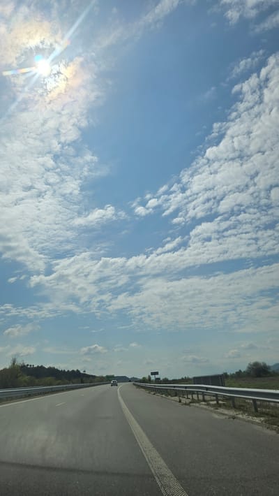 The image shows a highway with a clear blue sky filled with scattered white clouds. The sun is visible in the lower left corner, emitting bright rays. The highway has a shoulder with a metal guardrail. A single white line divides the lanes. In the distance, green trees line the horizon, and a vehicle is visible on the road. This scene is likely situated in the city of Valkovo, Bulgaria.