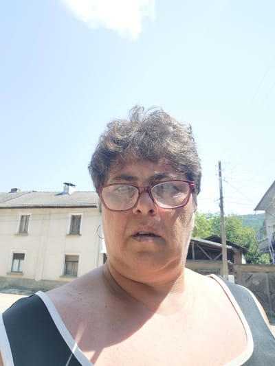 The image is a close-up, front-facing selfie of a woman with short, dark, curly hair and glasses. She is outdoors, with a bright blue sky and some clouds visible in the background. Behind her, a light-colored building with multiple windows and a tiled roof can be seen. Further in the background, there are trees, a wooden fence, and a utility pole. The woman is wearing a black and white tank top. Her skin appears to have some texture on her neck and chest. The scene is set in the city of Lomets, Bulgaria.