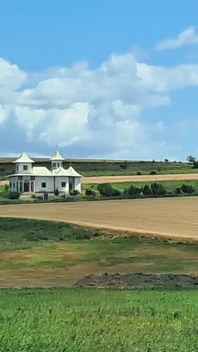The image depicts a white, ornate building with multiple small towers and a complex roofline, situated in a rural landscape. The building appears to be of a religious or historical nature, possibly a church or a manor house, with decorative elements and arched windows. It is surrounded by rolling hills and fields, with a clear blue sky dotted with fluffy white clouds above. The foreground features lush green vegetation, suggesting a natural, undeveloped setting. The distant background shows more fields and a treeline, contributing to the rural ambiance. The overall scene conveys a sense of peacefulness and isolation.