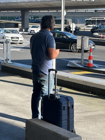 A medium shot, taken during daylight, shows a man with dark hair wearing a blue shirt and jeans standing next to a blue suitcase with a handle. He appears to be looking at something in his hand.
The location is Tremblay-en-France, France. A roadway with cars is visible in the background, along with an elevated structure and various signage. Traffic cones are present near the vehicles. The man and suitcase are positioned near a concrete structure, likely a part of the airport infrastructure.
