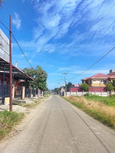 The image depicts a straight, paved road in Rhieng Mancang, Indonesia, under a clear blue sky with scattered white clouds. The road is flanked by residential properties. On the left side of the road, a single-story house with a dark red roof and a black metal fence is visible, partially obscured by a tall, rusty pole and overhead power lines. Adjacent to this house is a large, leafy green tree. Further down the road on the left, more houses and fences can be seen. On the right side of the road, a modern, two-story house with a prominent red roof and decorative yellow walls stands behind a low, ornate wall and a fence. This house has visible air conditioning units. The area along the road is characterized by dry grass and vegetation. Overhead power lines are a prominent feature throughout the scene, running parallel to the road. The image appears to be taken during daylight, likely in the morning or afternoon. No people are visible in the frame.