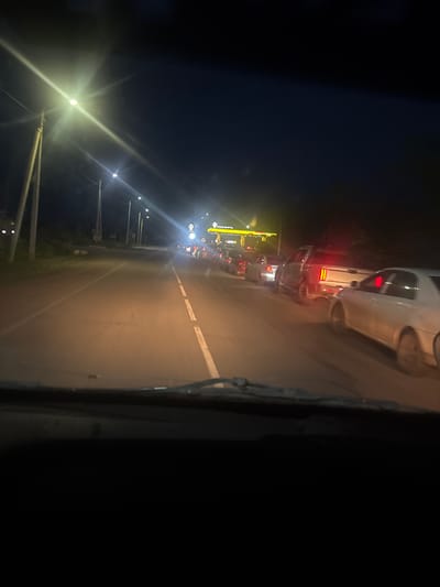 The image depicts a nighttime scene of a road in Petropavlovka, Russia, possibly at a gas station or service area. A queue of cars is visible, illuminated by various light sources. The time of day is clearly night, and the weather appears clear.

In the foreground, the camera is positioned inside a vehicle, looking out through the windshield, which has a wiper blade visible on the right side. The road surface is asphalt with white lane markings. To the left, there are streetlights and signs, one of which displays yellow lettering that reads "РОСПРОМБАНК". Further down the road, a bright yellow sign with red lettering is also visible, likely indicating a business.

The main activity is vehicles waiting in line, suggesting a stop at a service station, a traffic congestion point, or a toll booth. Several cars are lined up, their taillights and headlights casting reflections on the wet-looking road surface. The overall atmosphere is one of movement and waiting during nighttime hours. The lighting from the signs and streetlights creates a somewhat dramatic and high-contrast scene against the dark night sky. No people are clearly discernible.