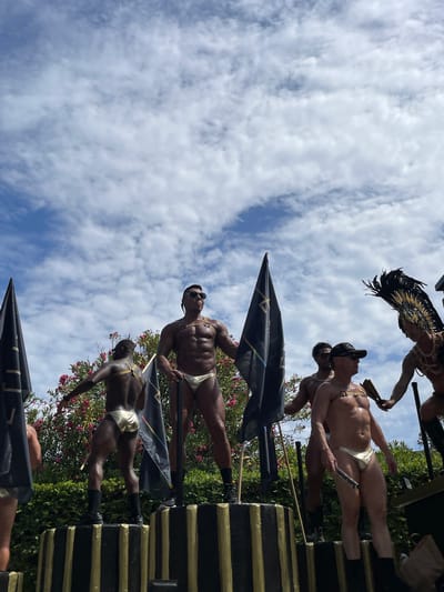 This image captures a scene from a parade in Sitges, Spain, likely during Pride celebrations. Several men are prominently featured, some wearing minimal, gold-colored attire such as briefs and harnesses. One individual wears an elaborate headdress with black feathers and gold accents, accessorized with jewelry. Another man holds a fan. They appear to be standing on a raised platform decorated with black and gold stripes. In the background, there are pink flowering bushes and green foliage. The sky above is partly cloudy with a bright blue expanse. Several large black flags with gold designs are also visible, held by participants or as part of the parade float. The overall atmosphere suggests a festive and celebratory event.